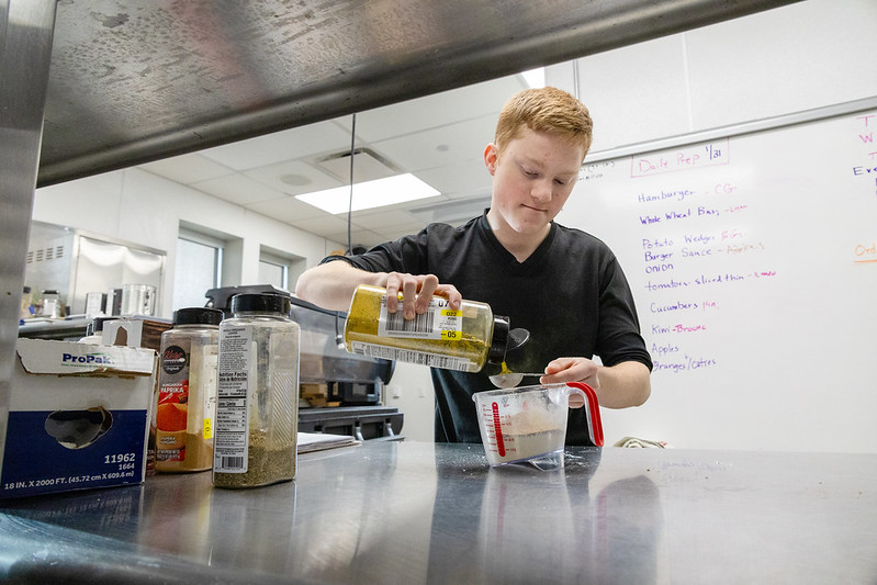 A redheaded teenager pouring a spice into a measuring spoon over a measuring cup. 