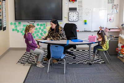 Elementary teacher at a table talking with one student while another student writes or draws