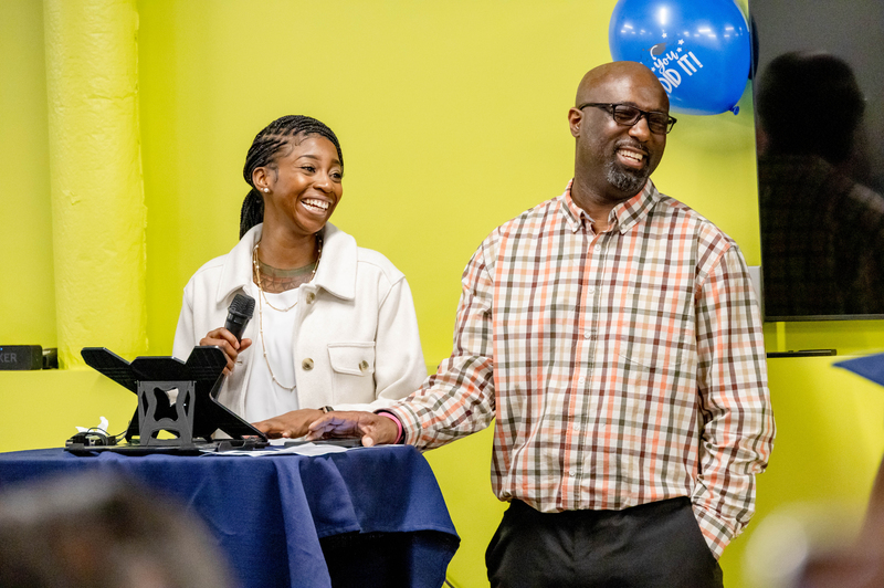 A woman and a man smile while standing at a podium. 