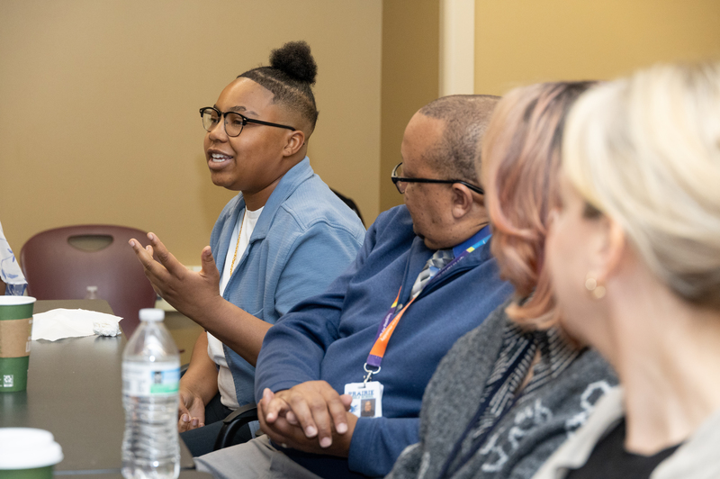A student apprentice shares her experience in the program so far while seated at a table with three people on her left looking at her.