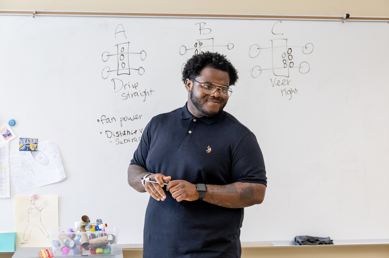 A student apprentice smiles while teaching class in front of a white board with formulas written on it. 